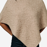 A woman wearing a beige knit poncho sweater with a textured geometric diamond pattern, paired with dark blue jeans, shown against a plain light gray background.
