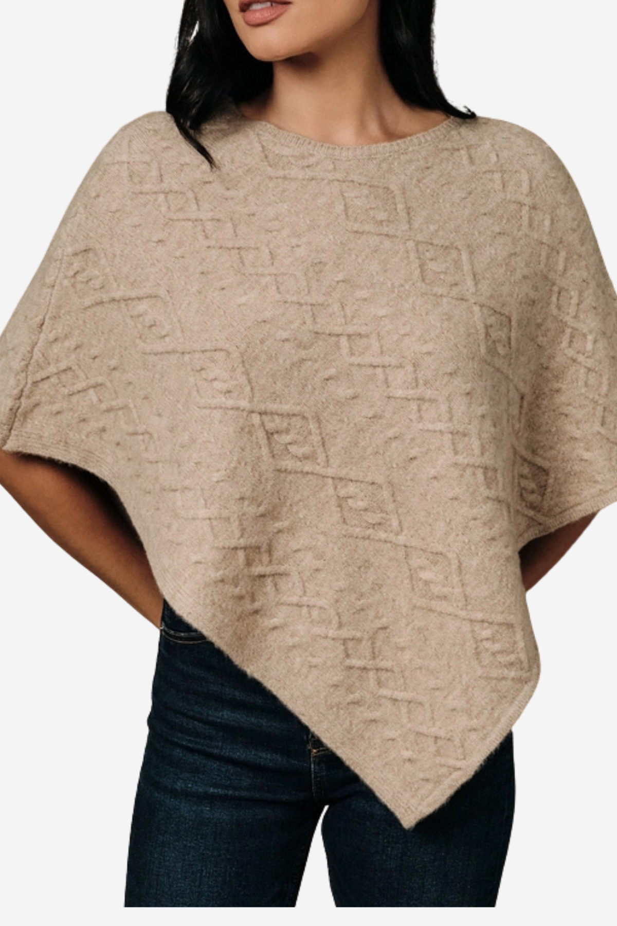 A woman wearing a beige knit poncho sweater with a textured geometric diamond pattern, paired with dark blue jeans, shown against a plain light gray background.