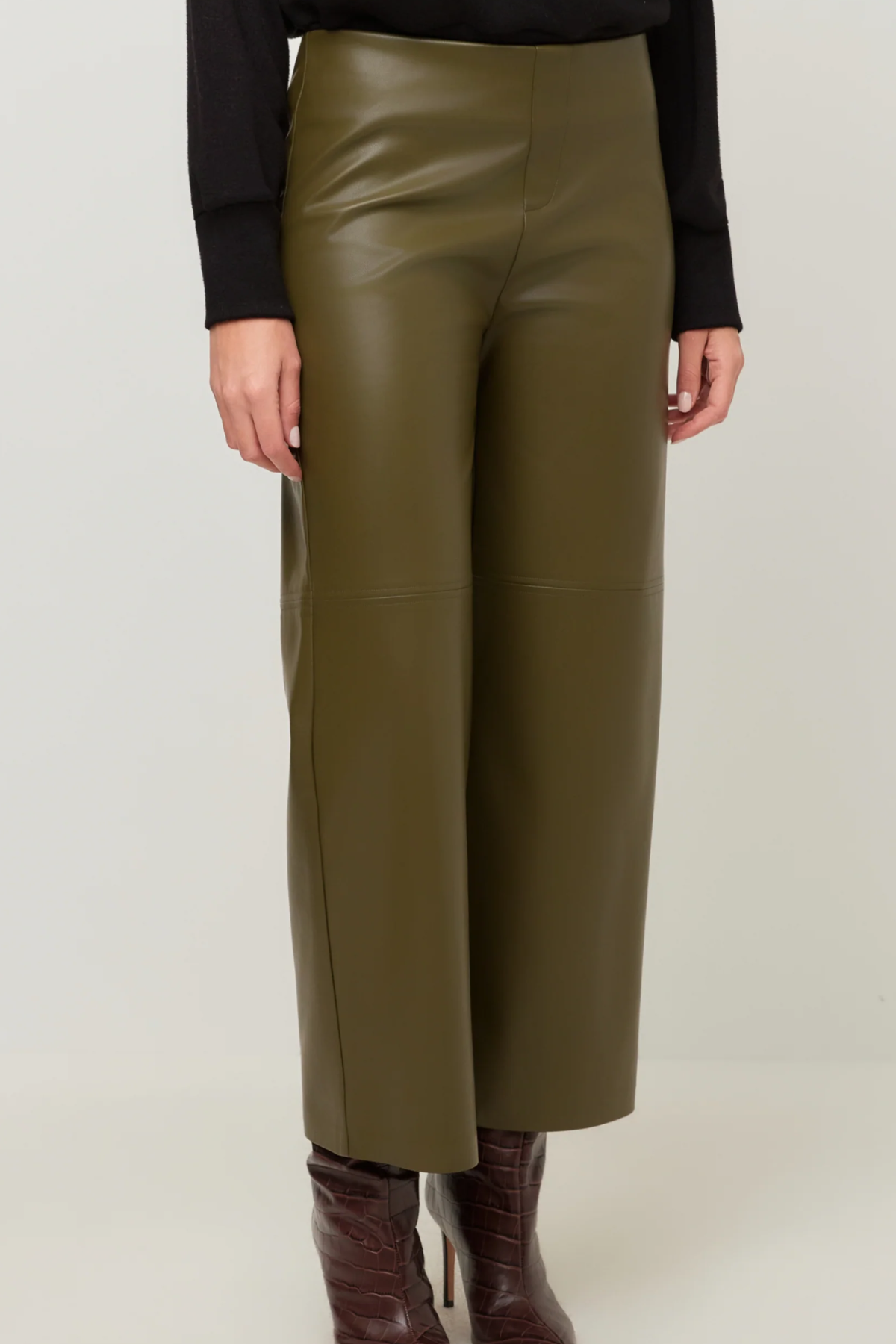 A person wearing olive green wide-leg faux leather pants, styled with a black long-sleeve top and brown leather ankle boots, against a light gray studio background.