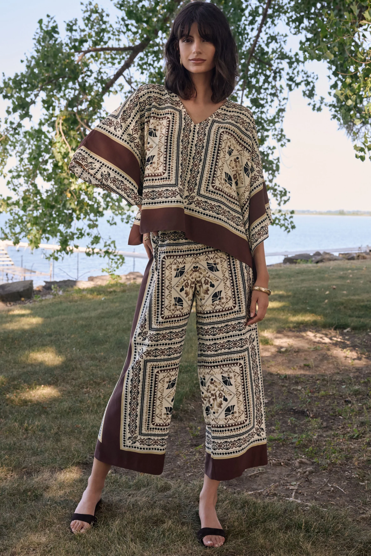Woman wearing a patterned outfit standing outdoors with trees and water in the background