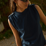 Woman wearing a dark blue sleeveless top and sunglasses, standing outdoors with greenery in the background.