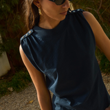 Woman wearing a dark blue sleeveless top and sunglasses, standing outdoors with greenery in the background.