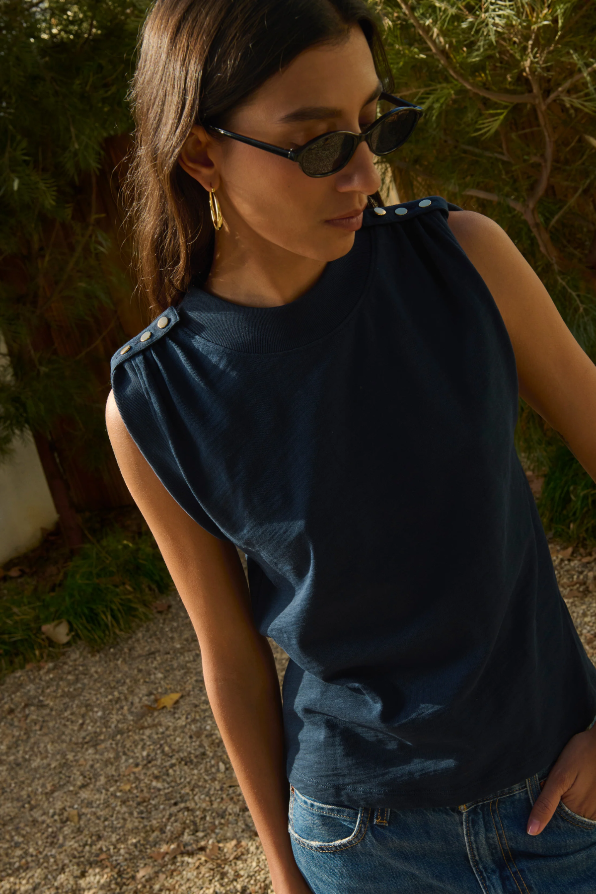 Woman wearing a dark blue sleeveless top and sunglasses, standing outdoors with greenery in the background.
