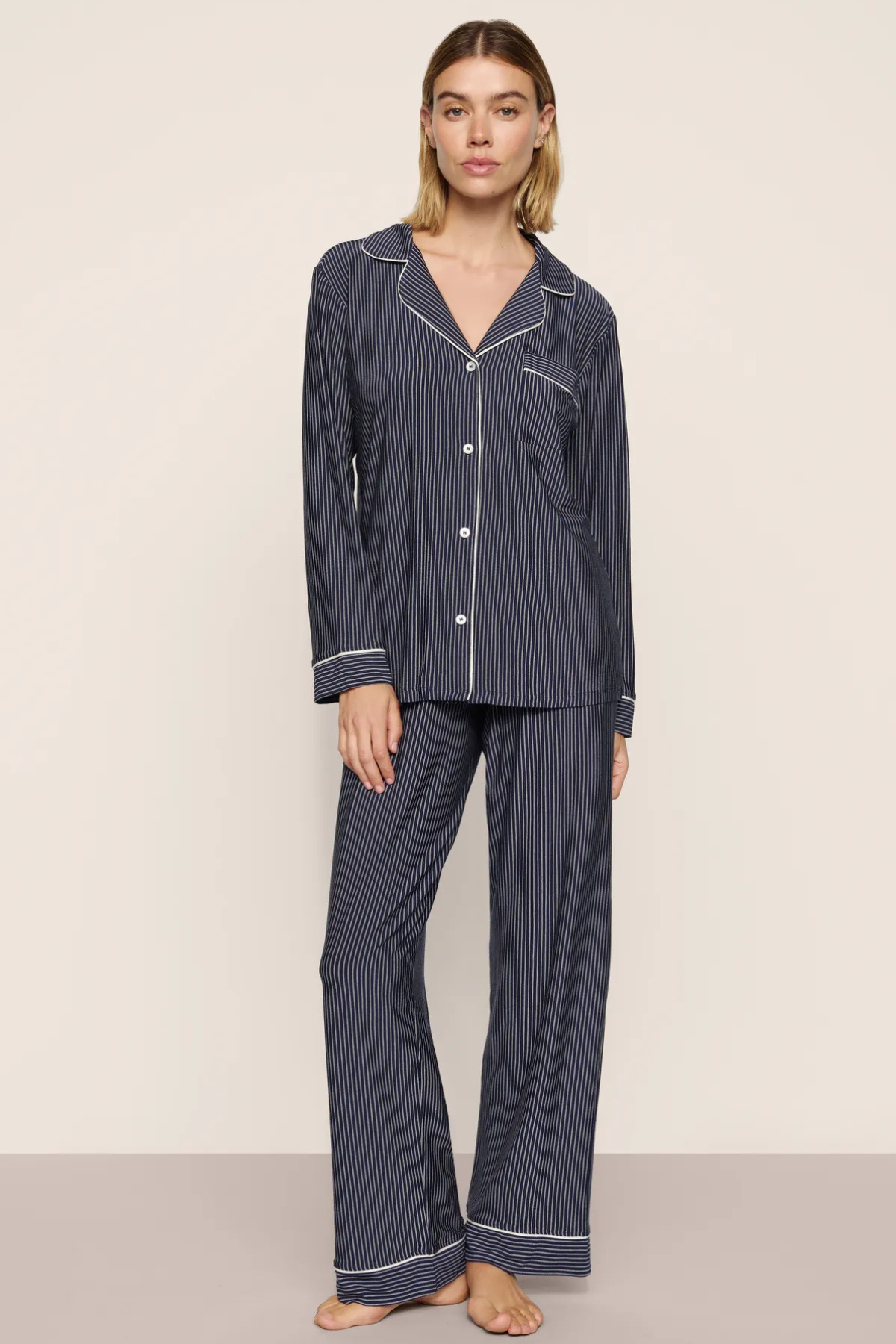 A woman modeling a two-piece pajama set with dark navy blue and white pinstripes, featuring a long-sleeve button-front top and matching wide-leg pants, set against a plain beige studio background.