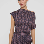 A woman modeling a dark purple off-the-shoulder jumpsuit with a pink and white geometric pattern, photographed against a light gray studio background.