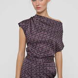 A woman modeling a dark purple off-the-shoulder jumpsuit with a pink and white geometric pattern, photographed against a light gray studio background.
