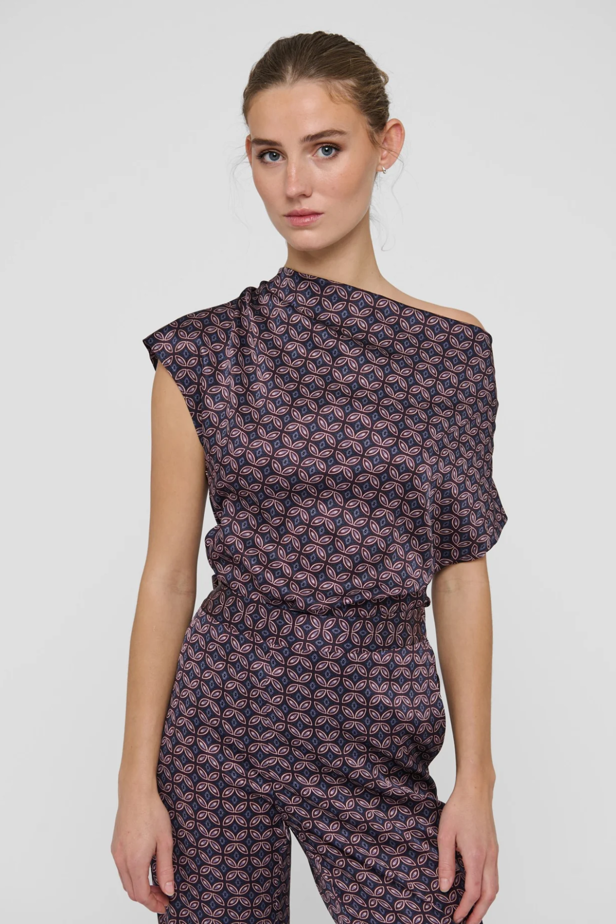 A woman modeling a dark purple off-the-shoulder jumpsuit with a pink and white geometric pattern, photographed against a light gray studio background.