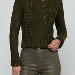A woman wearing a cropped, long-sleeve cable-knit sweater in olive green, paired with dark olive faux-leather pants, against a light gray studio background.