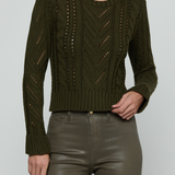A woman wearing a cropped, long-sleeve cable-knit sweater in olive green, paired with dark olive faux-leather pants, against a light gray studio background.