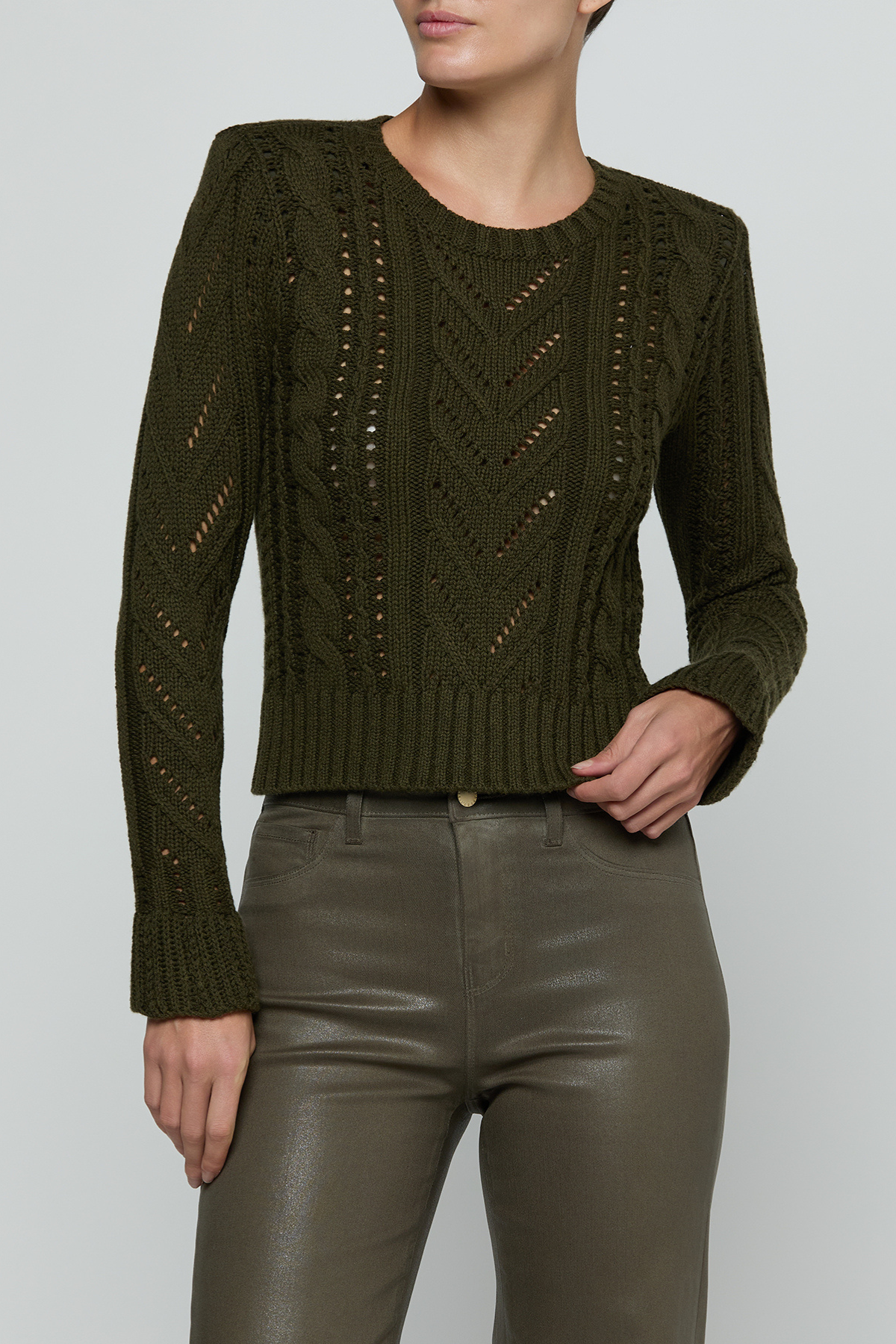 A woman wearing a cropped, long-sleeve cable-knit sweater in olive green, paired with dark olive faux-leather pants, against a light gray studio background.