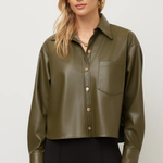 A woman modeling a cropped, long-sleeve olive green faux-leather button-up shirt with a chest pocket, paired with black pants, against a light gray studio background.