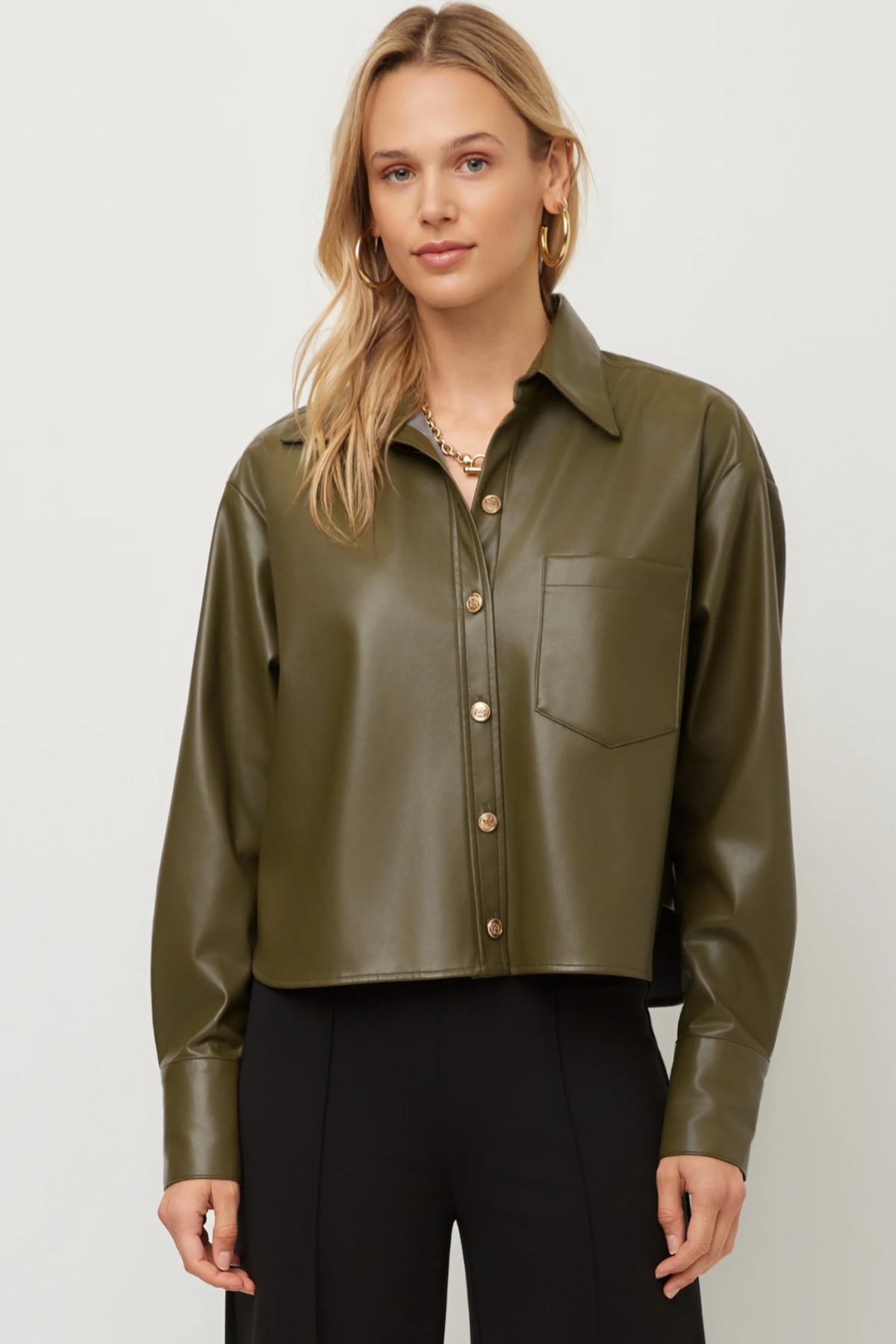 A woman modeling a cropped, long-sleeve olive green faux-leather button-up shirt with a chest pocket, paired with black pants, against a light gray studio background.