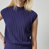 A woman wearing a sleeveless ribbed knit mock neck sweater in a deep purple color, paired with blue jeans, against a plain light gray background.
