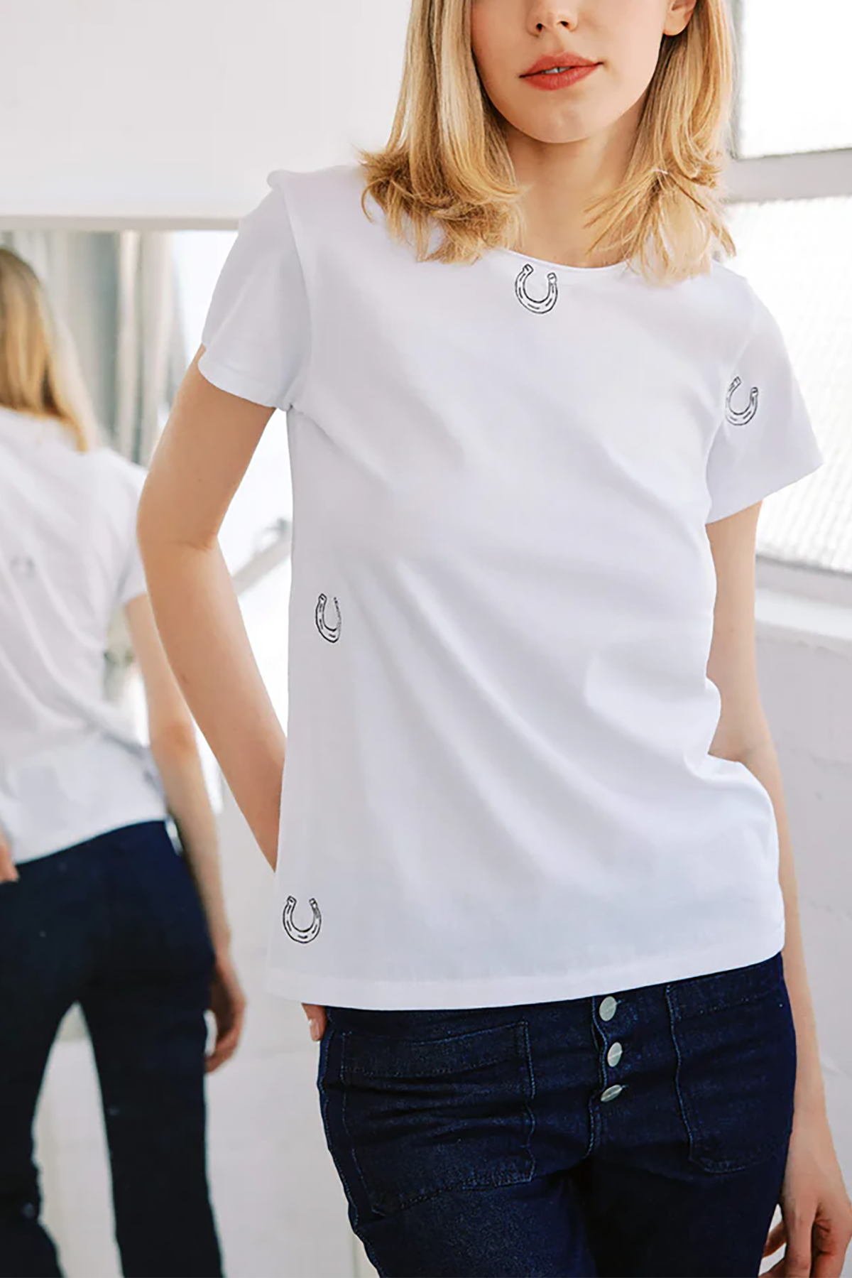 Woman wearing a white t-shirt with small logos, standing indoors.