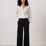 A woman stands in a studio wearing a white wrap-style blouse and black wide-leg trousers, with her hands in the trouser pockets. The background is a clean off-white wall with a light wood floor.