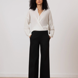 A woman stands in a studio wearing a white wrap-style blouse and black wide-leg trousers, with her hands in the trouser pockets. The background is a clean off-white wall with a light wood floor.