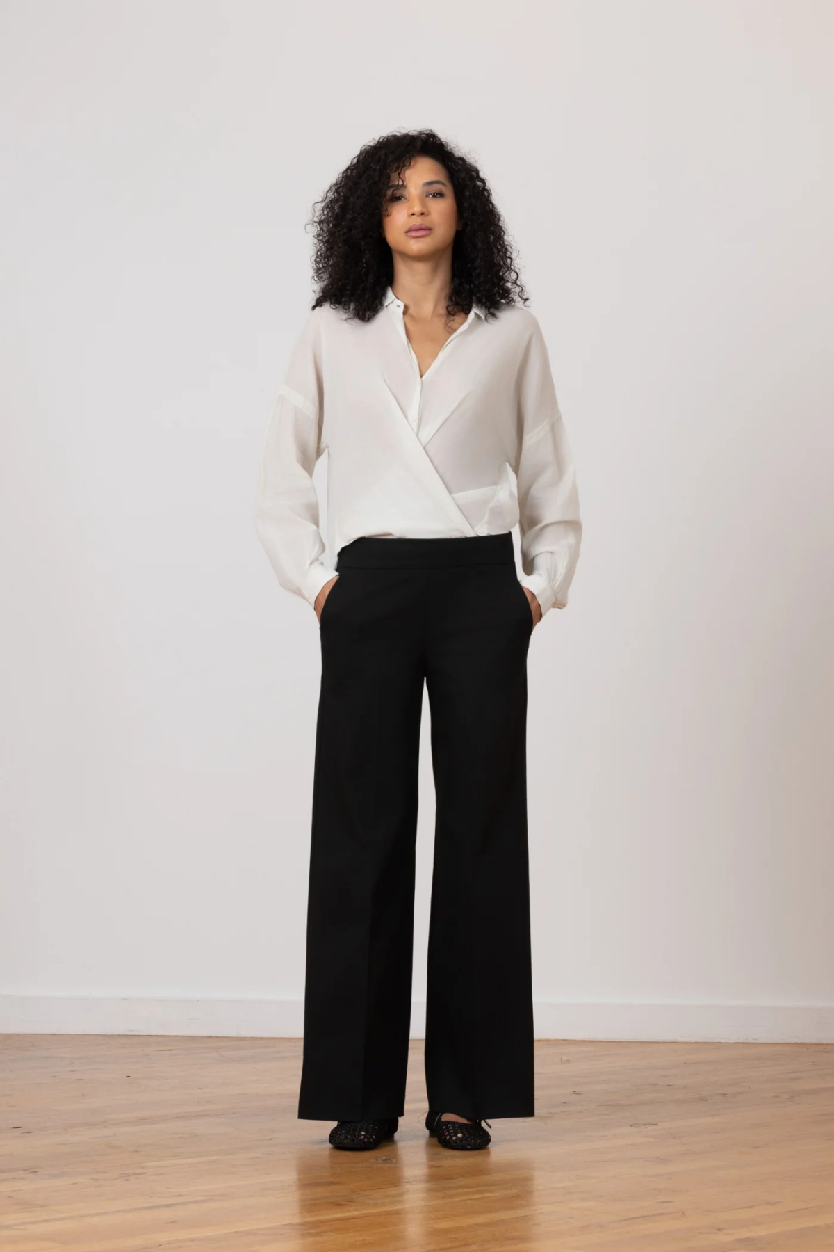 A woman stands in a studio wearing a white wrap-style blouse and black wide-leg trousers, with her hands in the trouser pockets. The background is a clean off-white wall with a light wood floor.