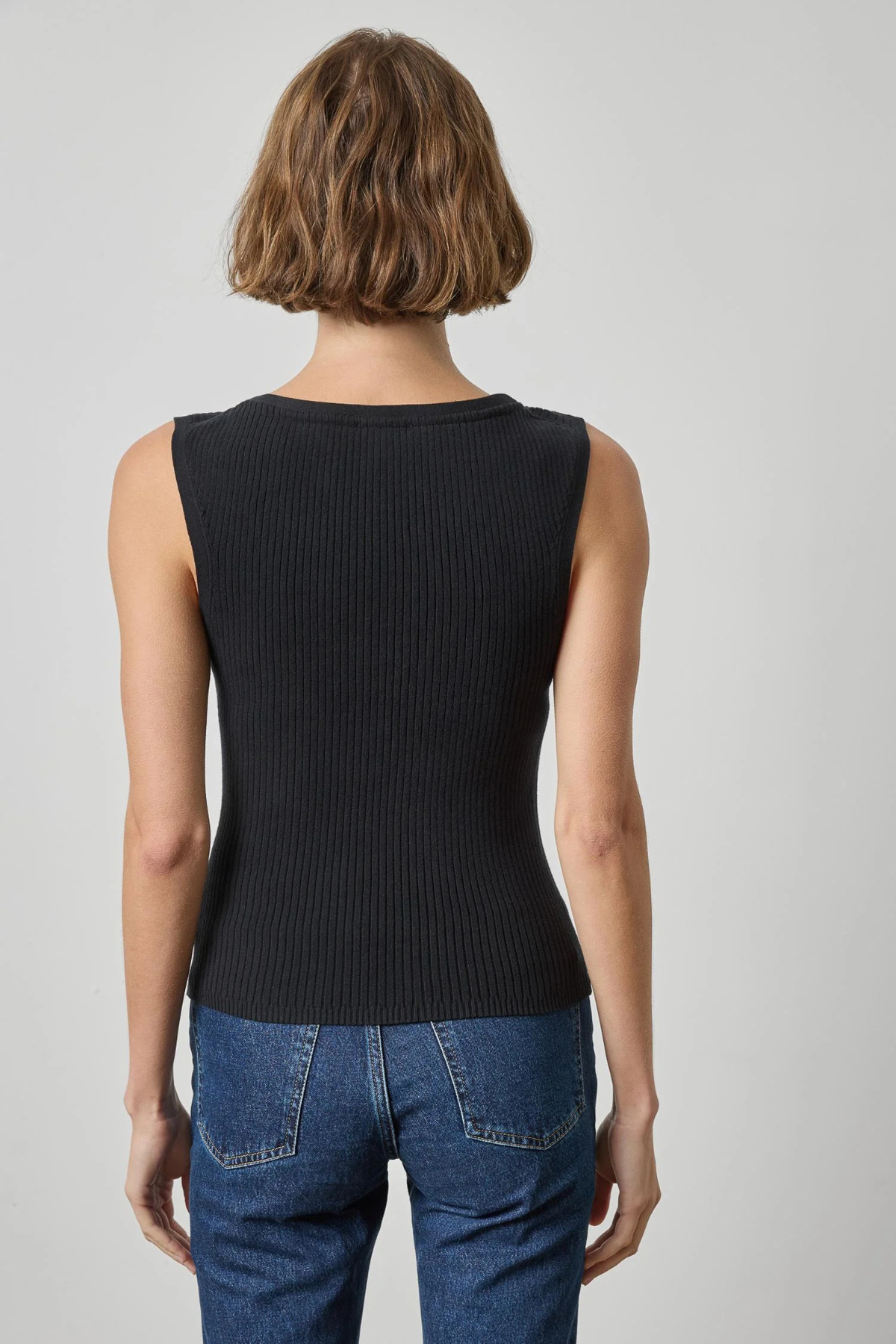 A woman viewed from the back wearing a sleeveless ribbed black top and blue jeans against a light gray studio background.