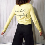 Person wearing a light yellow sweater with text and black shorts against a white paneled wall.