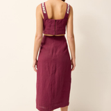 A woman shown from the back wearing a sleeveless two-piece linen set in deep burgundy, featuring a cropped top with embroidered straps and a matching midi skirt, paired with brown strappy sandals against a plain off-white background.