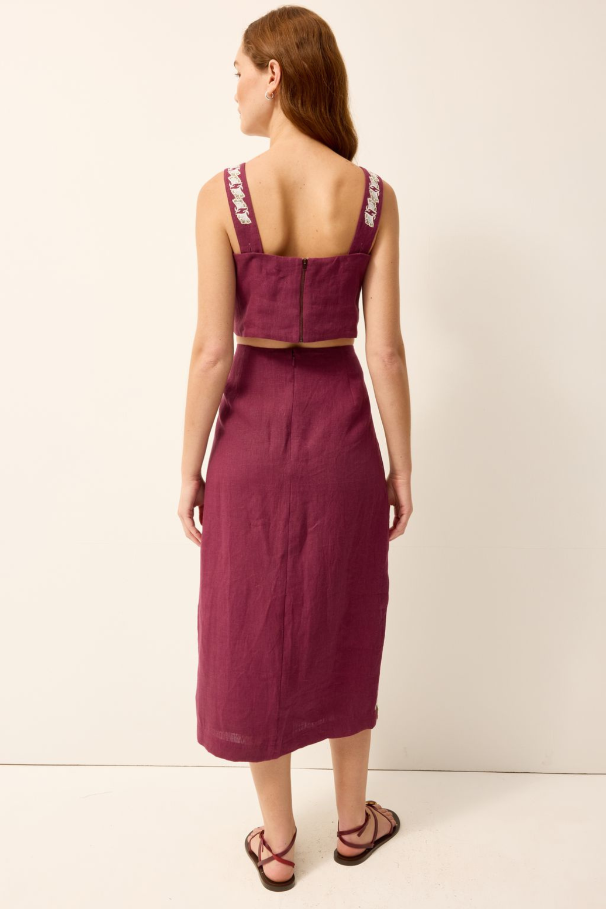 A woman shown from the back wearing a sleeveless two-piece linen set in deep burgundy, featuring a cropped top with embroidered straps and a matching midi skirt, paired with brown strappy sandals against a plain off-white background.