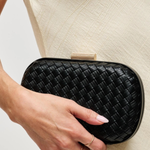 A close-up of a person holding a black woven leather clutch bag with a gold-tone clasp against a light beige textured dress.