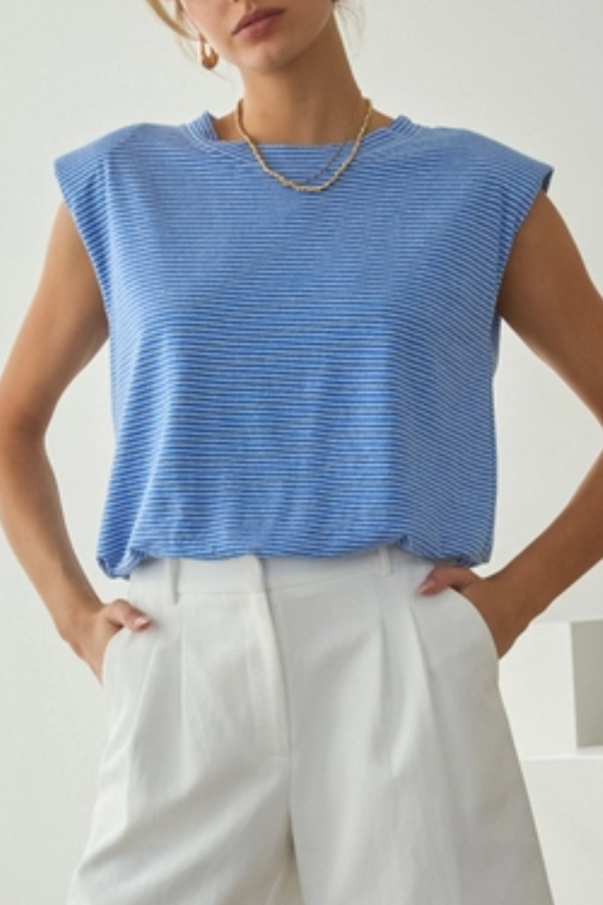 Woman wearing a blue sleeveless top and white pants against a plain background