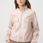 A woman wearing a light blush pink zip-up jacket with a stand collar and two front flap pockets, paired with taupe trousers, against a light gray studio background.