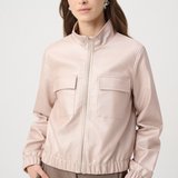 A woman wearing a light blush pink zip-up jacket with a stand collar and two front flap pockets, paired with taupe trousers, against a light gray studio background.