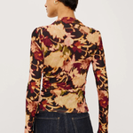 A woman shown from the back wearing a fitted long-sleeve top with an abstract floral or leaf-like print in beige, rust red, brown, and muted green on a black base, paired with dark blue jeans against a plain light background.