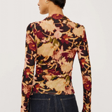 A woman shown from the back wearing a fitted long-sleeve top with an abstract floral or leaf-like print in beige, rust red, brown, and muted green on a black base, paired with dark blue jeans against a plain light background.