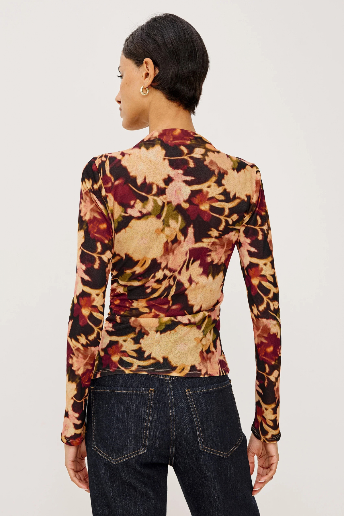 A woman shown from the back wearing a fitted long-sleeve top with an abstract floral or leaf-like print in beige, rust red, brown, and muted green on a black base, paired with dark blue jeans against a plain light background.