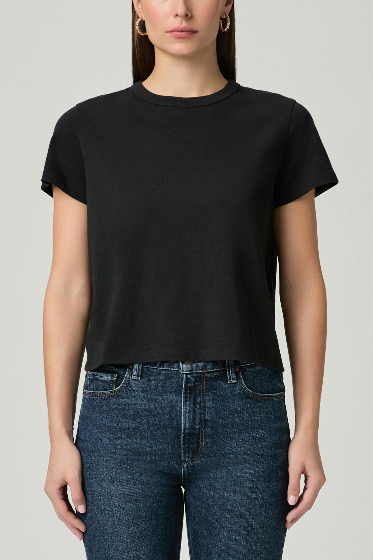 A woman wearing a black short-sleeve crewneck crop top and dark blue jeans, shown from the nose down against a light gray studio background.