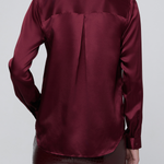 A person shown from the back wearing a long-sleeve satin blouse in deep burgundy with a subtle sheen, paired with a dark textured skirt, against a light gray studio background.
