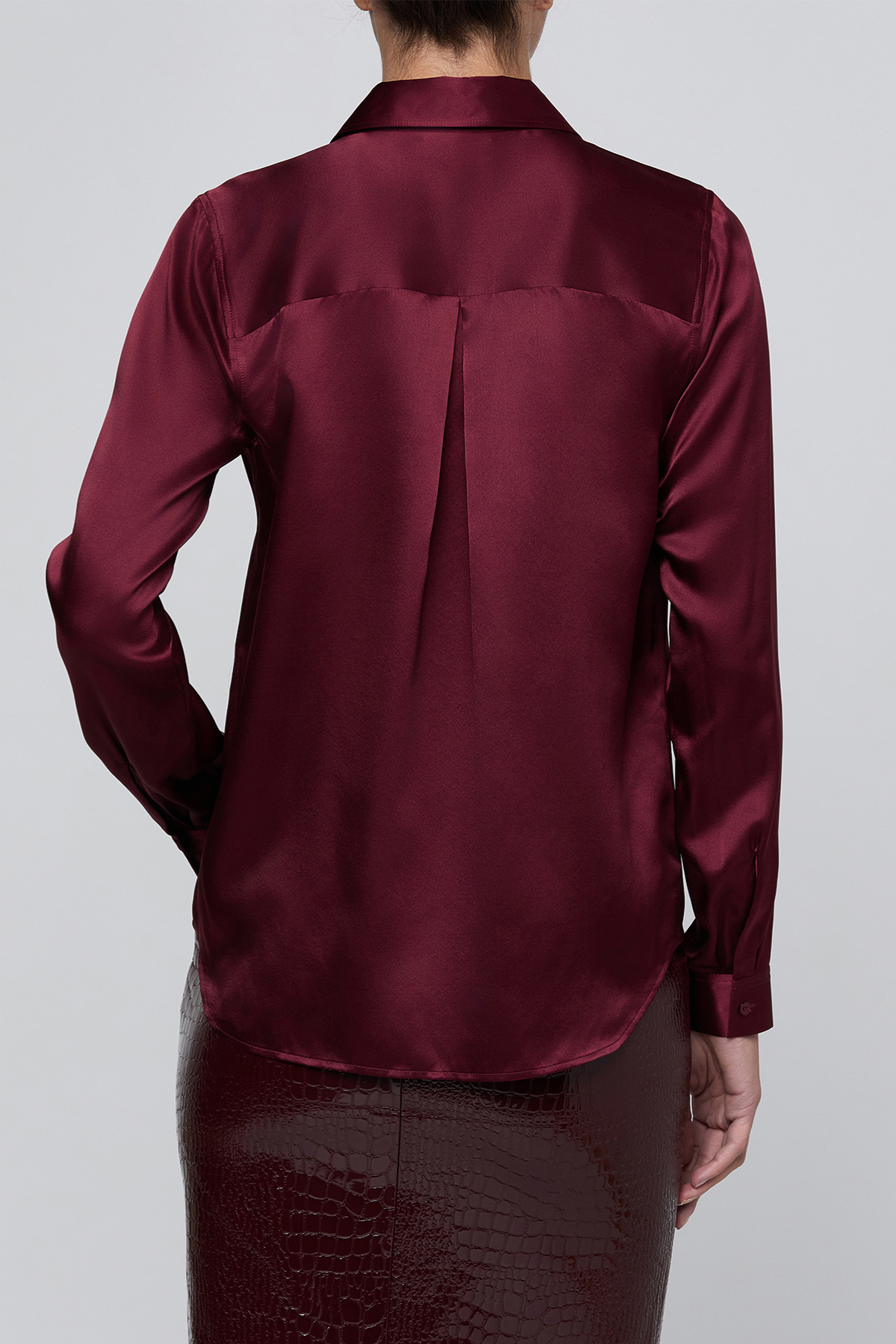 A person shown from the back wearing a long-sleeve satin blouse in deep burgundy with a subtle sheen, paired with a dark textured skirt, against a light gray studio background.