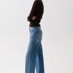 A woman modeling a pair of wide-leg blue jeans with silver chain detailing down the side, paired with a dark brown long-sleeve top and black pointed-toe heels against a plain white studio background.