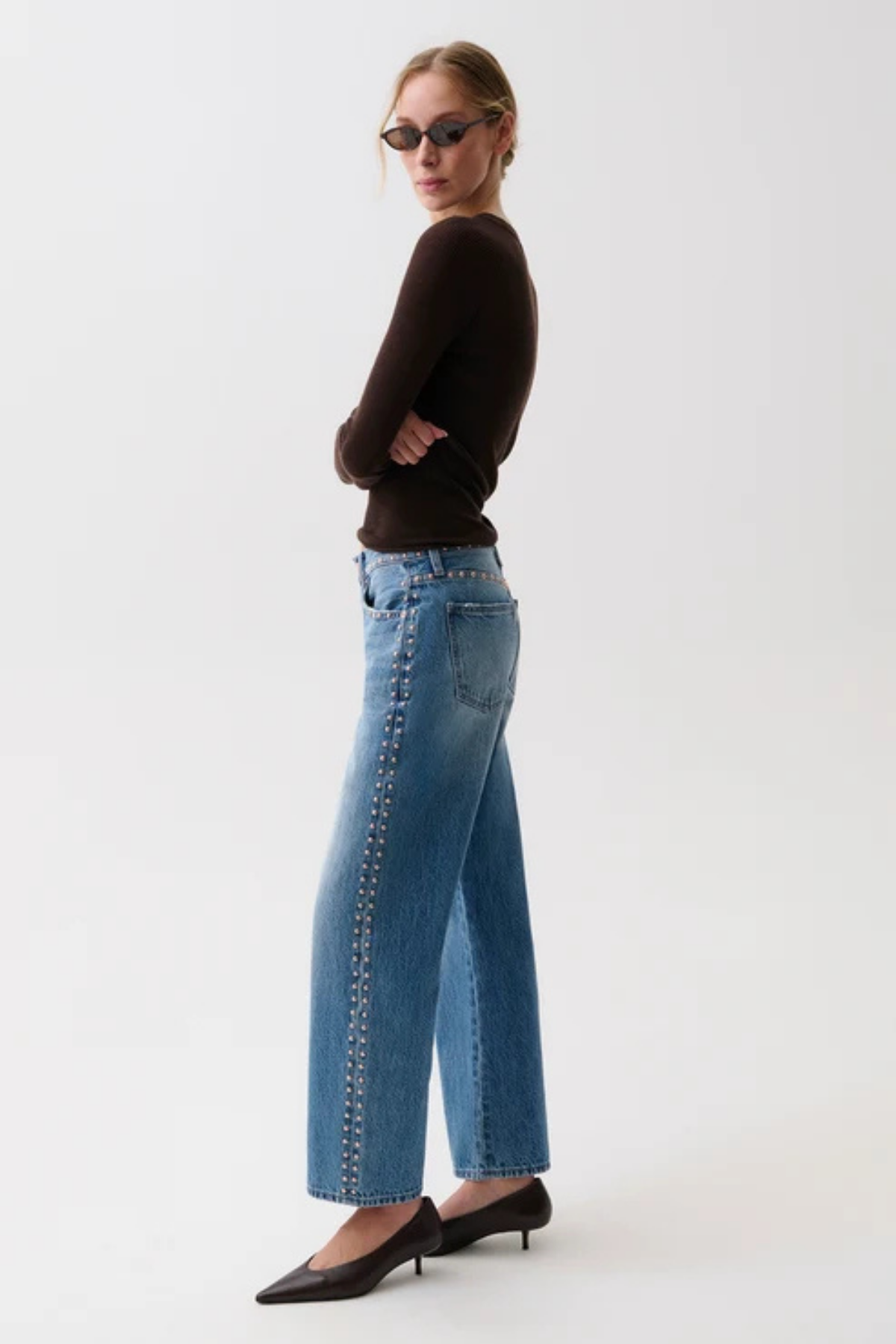 A woman modeling a pair of wide-leg blue jeans with silver chain detailing down the side, paired with a dark brown long-sleeve top and black pointed-toe heels against a plain white studio background.