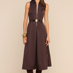 A woman modeling a sleeveless dark brown midi dress with a front zipper, a matching dark brown belt with a gold buckle, sunglasses, gold bracelets, and dark brown suede boots against a solid beige background.