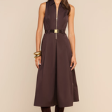 A woman modeling a sleeveless dark brown midi dress with a front zipper, a matching dark brown belt with a gold buckle, sunglasses, gold bracelets, and dark brown suede boots against a solid beige background.