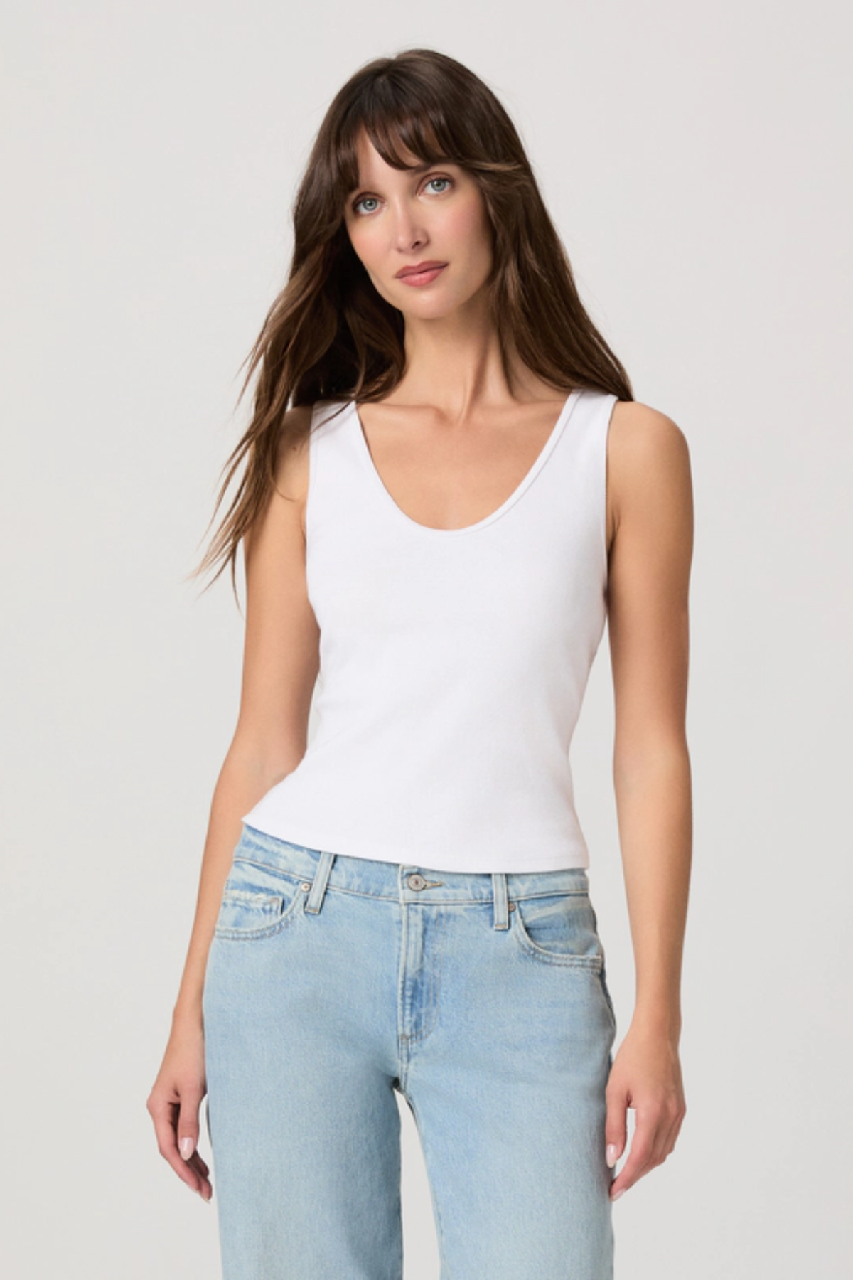 Woman wearing a white tank top and light blue jeans against a plain background