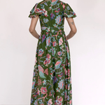 A woman viewed from the back wearing a green midi dress with a colorful floral and bird print, short flutter sleeves, and a gathered waist, paired with dark flat shoes against a plain white studio background.