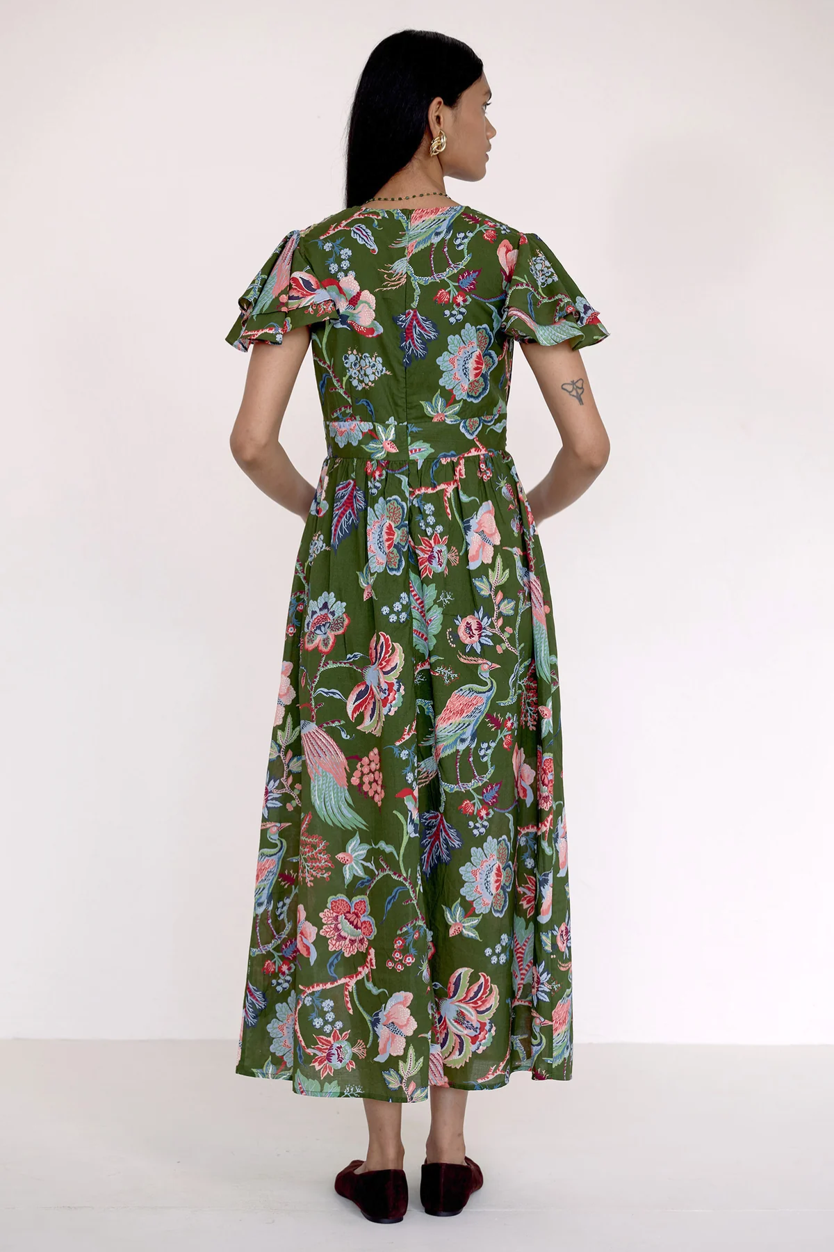 A woman viewed from the back wearing a green midi dress with a colorful floral and bird print, short flutter sleeves, and a gathered waist, paired with dark flat shoes against a plain white studio background.