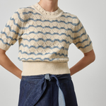 A woman wearing a cream short-sleeve knit sweater with blue wavy stripe patterns and a matching ribbed hem, paired with a dark blue denim skirt, against a light gray studio background.