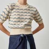 A woman wearing a cream short-sleeve knit sweater with blue wavy stripe patterns and a matching ribbed hem, paired with a dark blue denim skirt, against a light gray studio background.