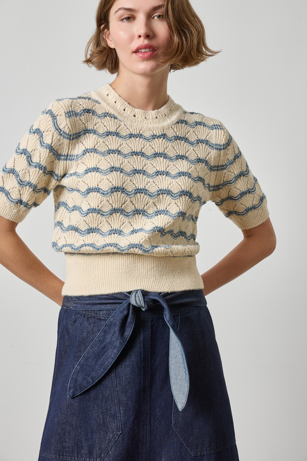 A woman wearing a cream short-sleeve knit sweater with blue wavy stripe patterns and a matching ribbed hem, paired with a dark blue denim skirt, against a light gray studio background.
