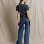 A woman stands with her back to the camera wearing a dark charcoal gray t-shirt and wide-leg blue denim jeans, posed against a draped beige fabric backdrop.