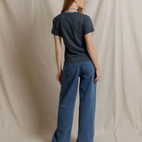 A woman stands with her back to the camera wearing a dark charcoal gray t-shirt and wide-leg blue denim jeans, posed against a draped beige fabric backdrop.
