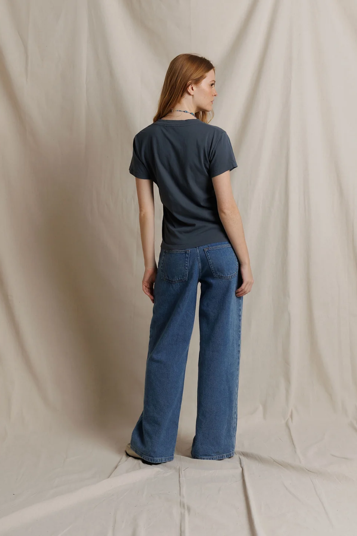 A woman stands with her back to the camera wearing a dark charcoal gray t-shirt and wide-leg blue denim jeans, posed against a draped beige fabric backdrop.