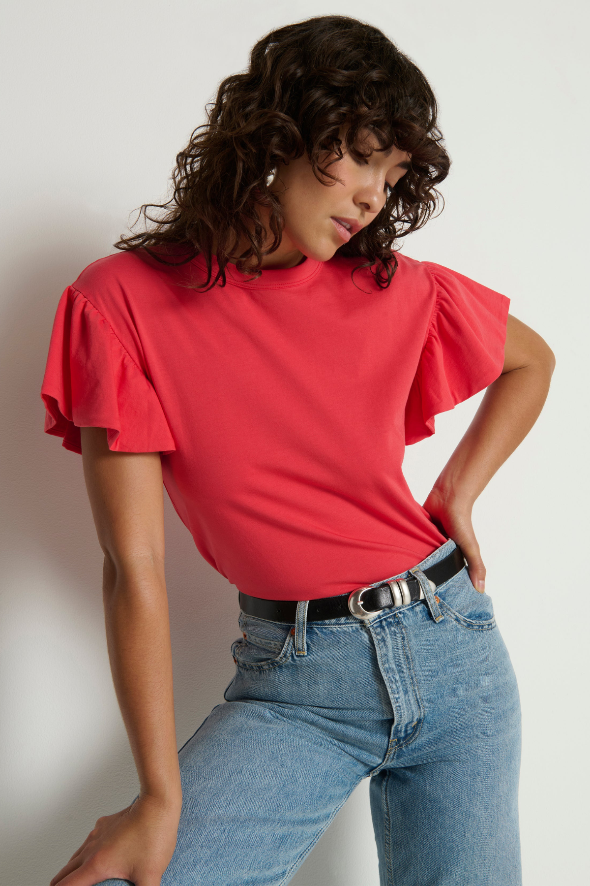 Woman wearing a red blouse with ruffled sleeves and blue jeans against a plain background