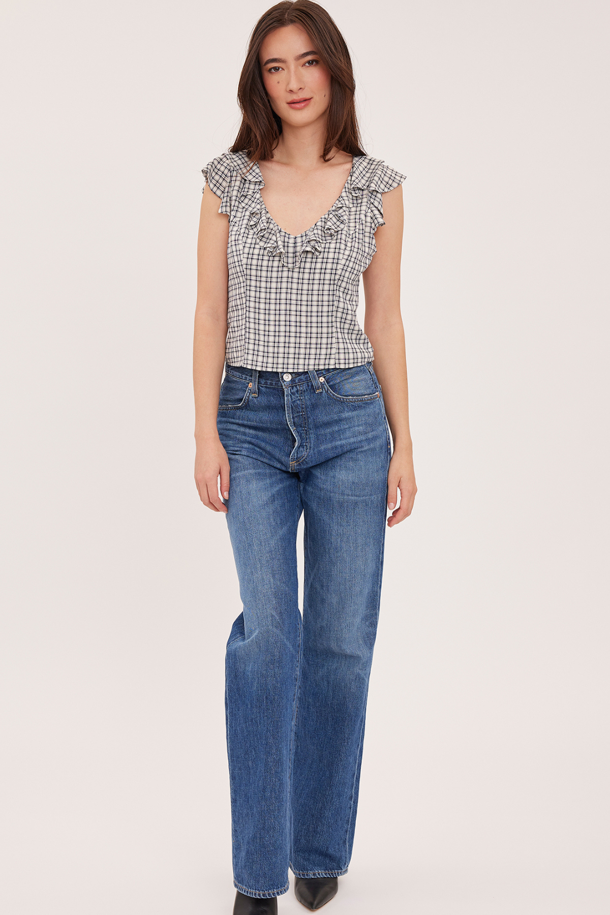 A woman modeling a sleeveless black and white gingham top with ruffle trim and blue wide-leg jeans against a plain light background.